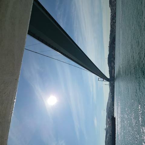       A long suspension bridge stretches across the Bosphorus with bright sun and blue sky overhead.
  