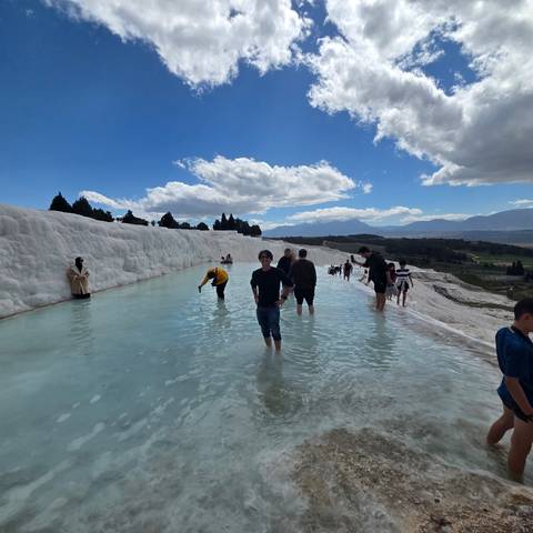       Visitors wade through the turquoise thermal pools of Pamukkale’s white travertine terraces under a partly cloudy sky.
  