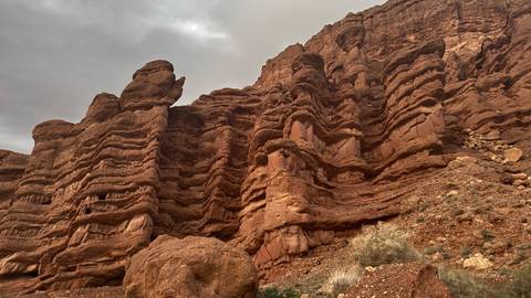       Towering layered red rock cliffs rise toward a cloudy sky in a rugged canyon landscape.
  