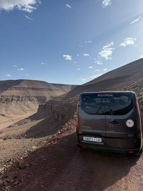       A tour van parks at a high desert overlook with steep eroded cliffs and a winding dirt road below.
  