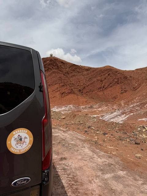       A tour van with a Morocco logo partly blocks a view of rust-colored desert hills.
  