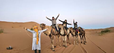       A caravan of tourists rides camels across soft desert sand under a pale sky with the moon visible.
  