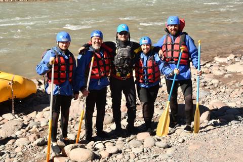       Five rafters in helmets and life jackets pose with paddles beside a fast-flowing river
  
