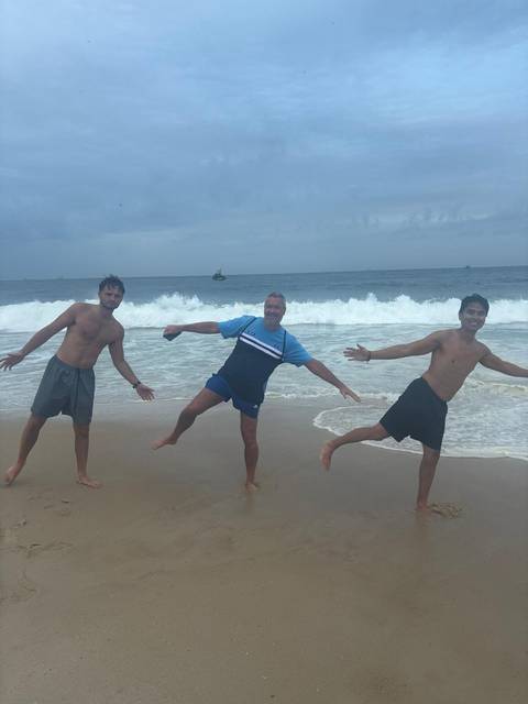       Three friends strike playful poses on a sandy beach with breaking surf and cloudy sky
  
