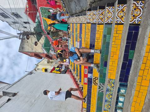       Traveller sits on colorful tiled steps of Selaron staircase surrounded by other visitors
  