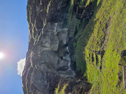       Rocky cave entrance cut into cliff face with grassy foreground steps and sun overhead
  