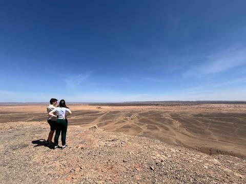       Couple stand on cliff edge overlooking vast desert plain under blue sky.
  