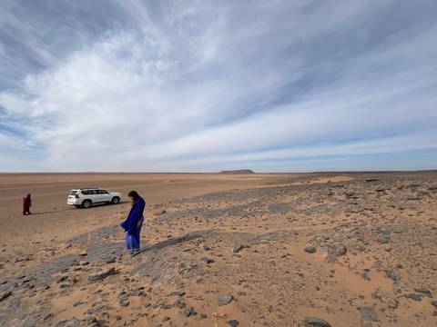       Sparse desert landscape with SUV and two travelers dressed in blue robes.
  