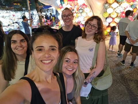       Close-up selfie of five friends in a bustling market surrounded by colorful lights and stalls.
  