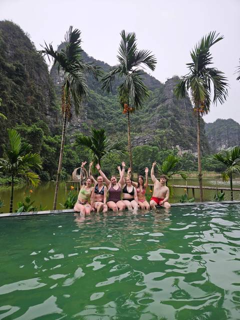       Group of friends in swimwear cheering from an infinity pool with limestone cliffs and lush forest behind.
  