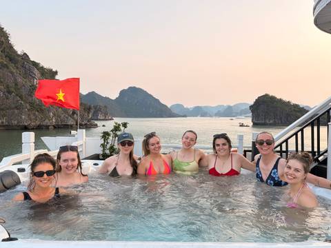       Eight travellers relaxing in a rooftop jacuzzi on a cruise ship with Halong Bay islands at sunset and a Vietnamese flag fluttering.
  