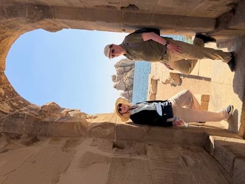       Two mature travellers posing under a stone archway overlooking blue water and granite formations.
  