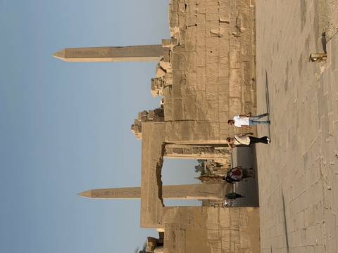       Visitors walk through a sandstone gateway flanked by towering obelisks and ruined temple walls under a clear blue sky.
  