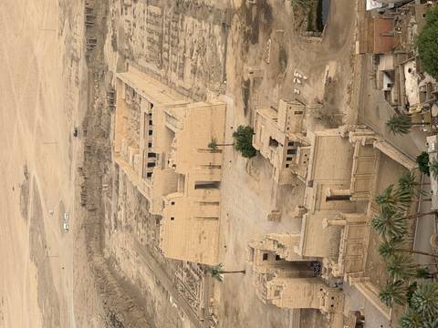       Aerial view of an ancient sandstone temple complex and surrounding desert landscape with palm trees at the edge.
  