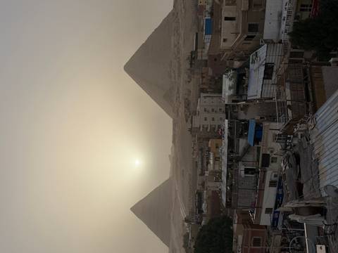       Sun setting through dusty haze over Giza with two pyramids silhouetted behind a foreground of rooftops.
  