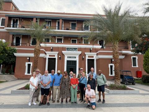       Tour group stands in front of a colonial-style hotel entrance framed by palm trees and landscaped gardens.
  