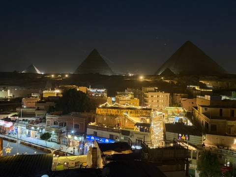       Night panorama of illuminated Giza pyramids towering over a lively bazaar-lit neighborhood.
  