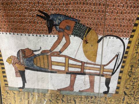       Wall painting shows Anubis performing embalming ritual on a mummy with rich colors and geometric background.
  