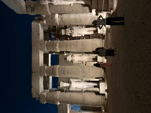       Illuminated statues and columns of Luxor Temple courtyard at night with small clusters of visitors.
  