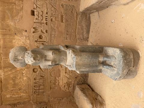       Granite statue of the lioness goddess Sekhmet seated against a wall of hieroglyphs in a sandy courtyard.
  
