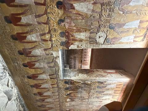       Colorful wall murals in an ancient tomb corridor depict rows of male figures in white kilts.
  
