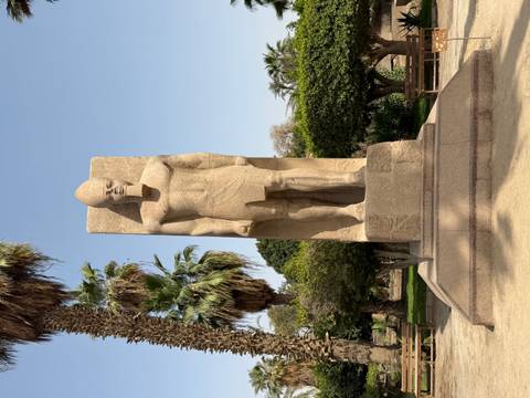       Granite statue of a pharaoh standing in a landscaped garden with palm trees and manicured hedges.
  