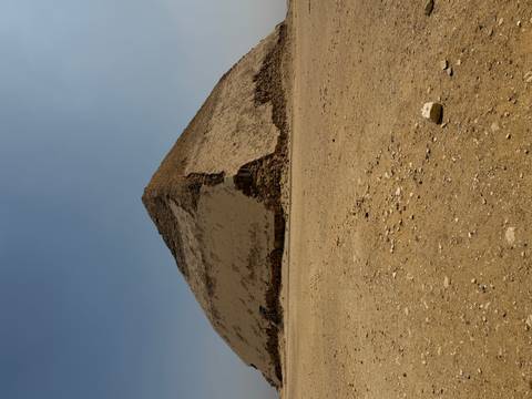       The Bent Pyramid shows its distinctive change in angle against a pale desert sky with rubble in the foreground.
  