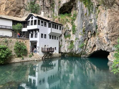       White dervish lodge nestled beside turquoise spring emerging from sheer rock cliff.
  