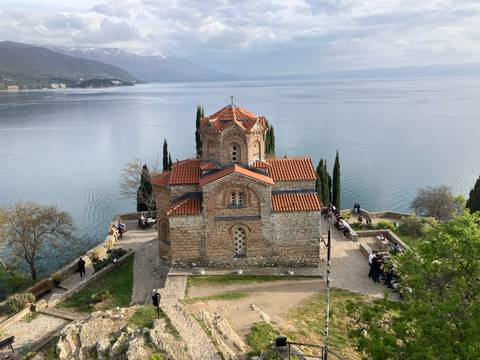       Byzantine stone church perched above shimmering lake with mountains on horizon.
  