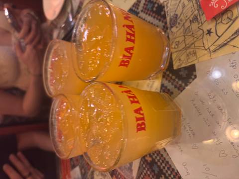      Four glasses of orange beverage marked 'Bia Ha Noi' on a crowded table.
  
