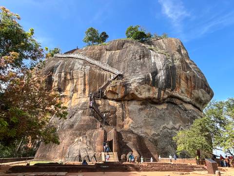       Massive monolithic Sigiriya rock fortress with zigzag staircases rises against blue sky.
  