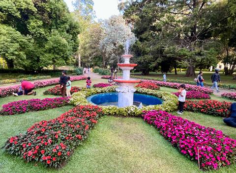       Colorful garden with central fountain surrounded by vibrant flowerbeds and tourists.
  