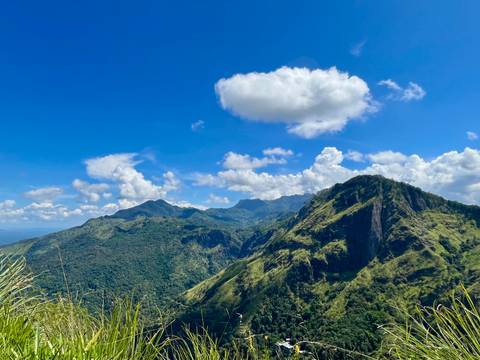       Panoramic view of lush green mountains beneath a bright blue sky with scattered clouds.
  