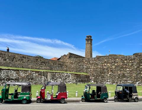       Stone ramparts and clock tower of Galle Fort with tuk tuks parked below.
  