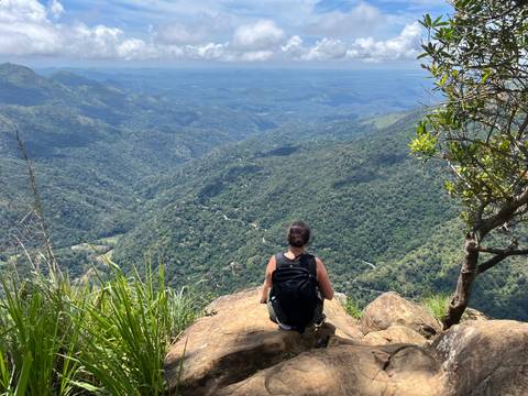       Traveler with backpack sits on a rocky overlook facing rolling green valleys.
  
