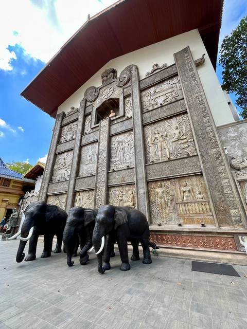       Stone relief wall adorned with Buddhist carvings and three elephant statues in front.
  