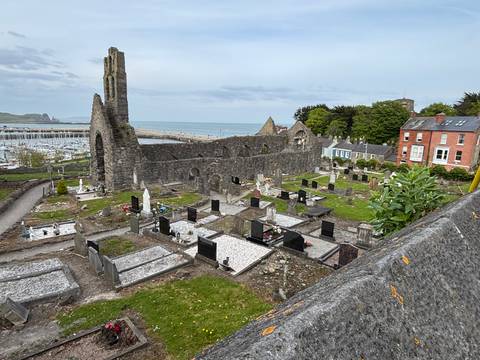       Ruined stone abbey and cliff-edge cemetery overlooking a harbour and open sea.
  