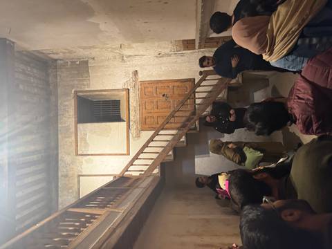       Visitors listen to a guide inside an aged stone building with wooden stairs and faded walls.
  