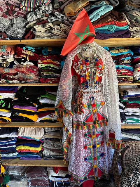       Colorful traditional Berber dress adorned with jewelry displayed in front of neatly stacked fabrics.
  