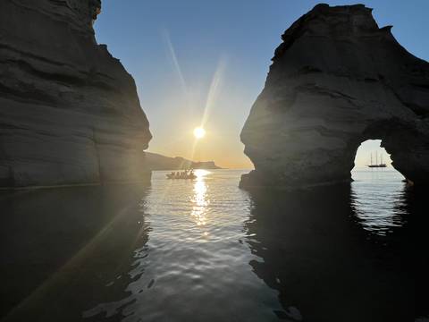       Sun gleaming through a natural sea arch with small boat and anchored ship nearby
  