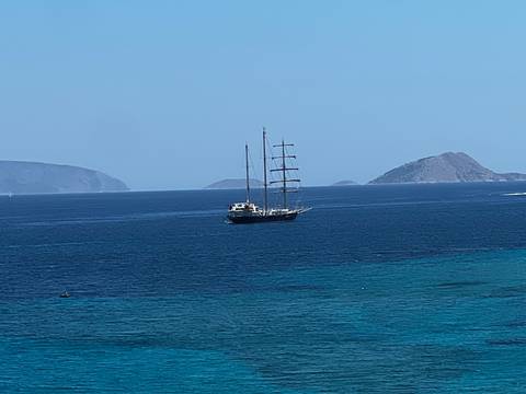       Distant view of a masted yacht anchored in deep blue Aegean waters with islands behind
  