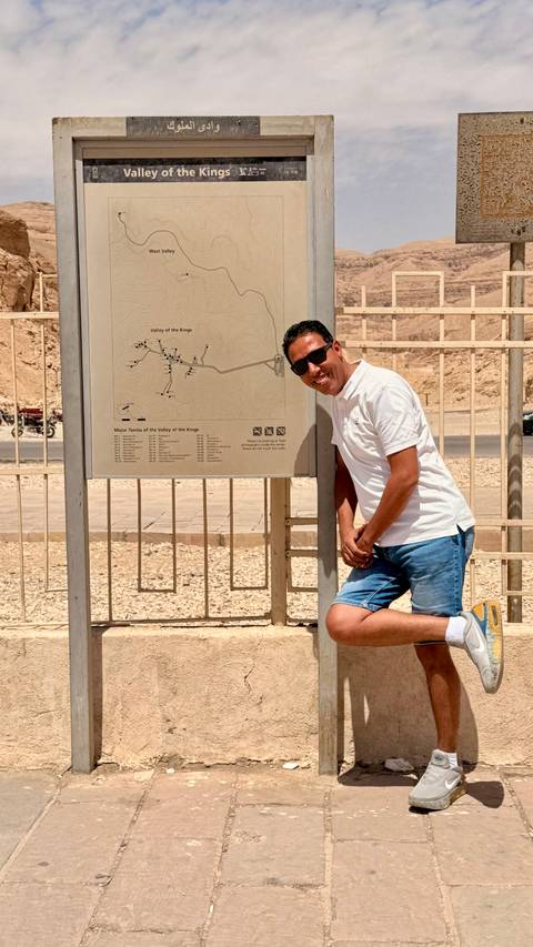       Traveler in white polo smiles beside an information map for the Valley of the Kings in a desert setting.
  