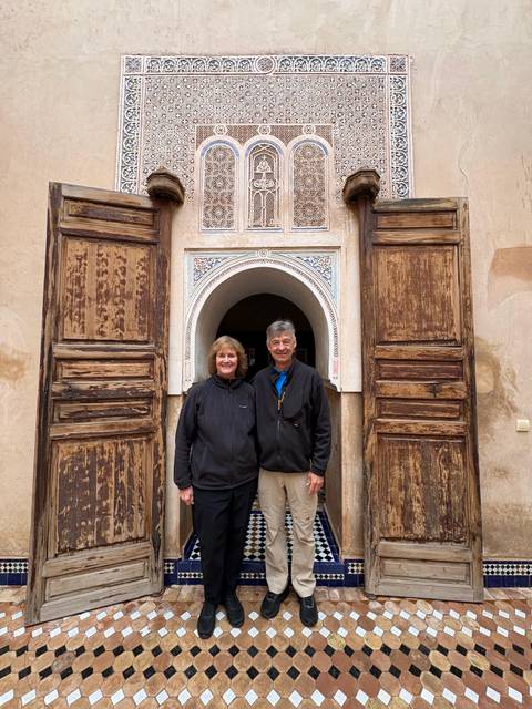       Smiling couple poses in front of an ornate wooden doorway with traditional Moroccan tilework.
  