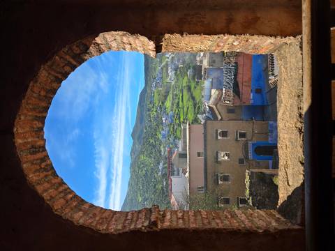       View through an arched stone window overlooking green hills and blue-washed houses of Chefchaouen.
  