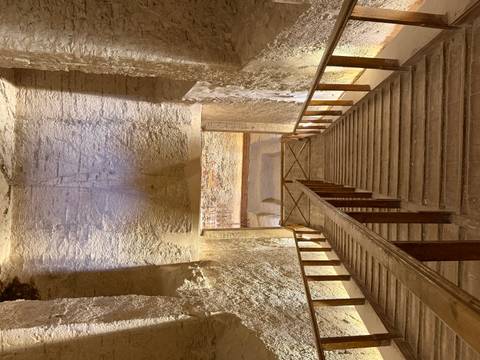       Dimly lit wooden staircase descending into the interior of an ancient Egyptian tomb with carved walls.
  