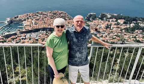       Smiling couple poses on a viewpoint railing high above Dubrovnik’s walled city and shimmering sea.
  