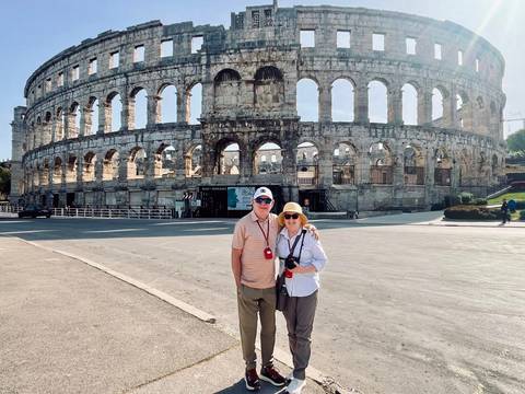       Couple standing in front of Pula’s Roman amphitheatre on a bright day, cameras ready.
  
