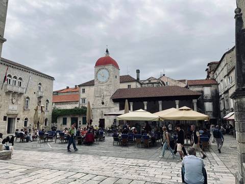       Lively town square in Trogir with a red-topped clock tower and café umbrellas.
  