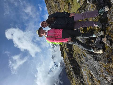       Two hikers smile on a rocky slope with glaciated Andean mountains rising into the clouds.
  