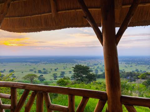       View across open plains from a wooden lodge balcony at sunrise with soft pastel skies.
  